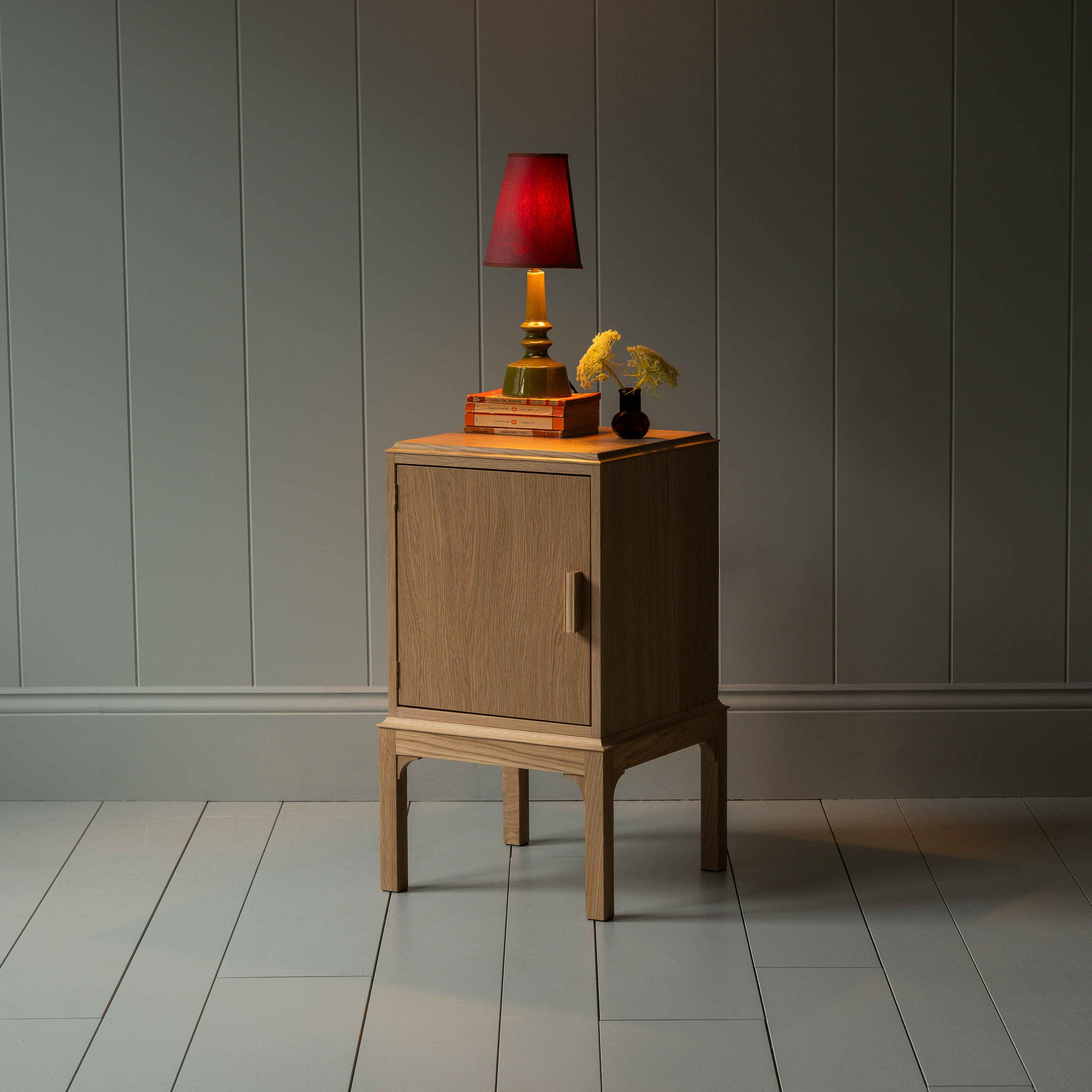 Bits and Bobs Bedside Table, Natural Oak, Left Hand Side Decorated With a table lamp and a small bottle of flowers - Nicola Harding