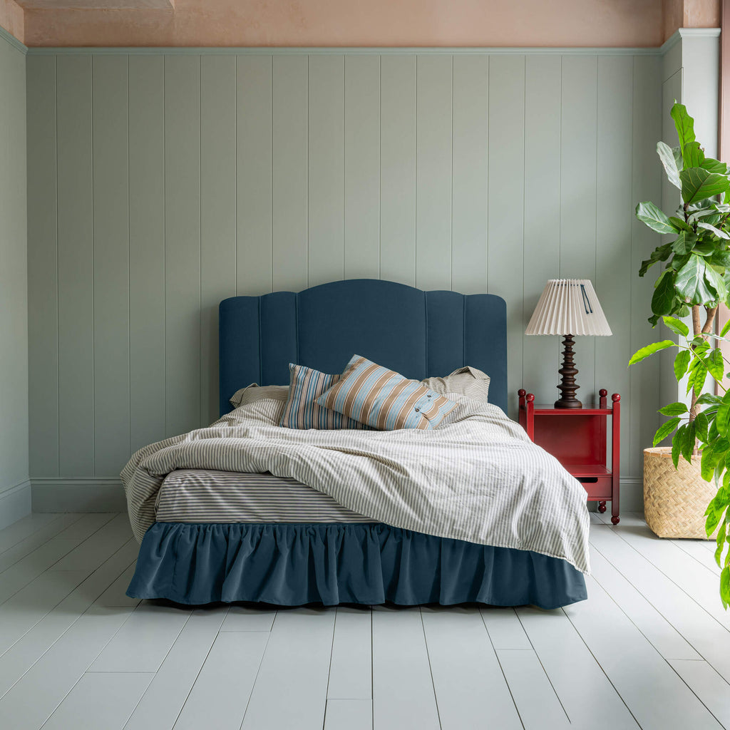 Cloud Nine Upholstered Bed in Intelligent Velvet Aegean with Ruffled Valance, accompanied by a red side table and a lamp, pillows, and a blanket.