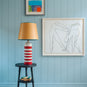 Humbug Straight Empire Paper Lamp Shade in Mustard with Blue Trim, With Humbug Striped Ceramic Table Lamp Base in Cherry Red & Warm White, On a Spindle Side Table, Carbon Black, With Beautiful Art Picture in the BackGround - Nicola Harding