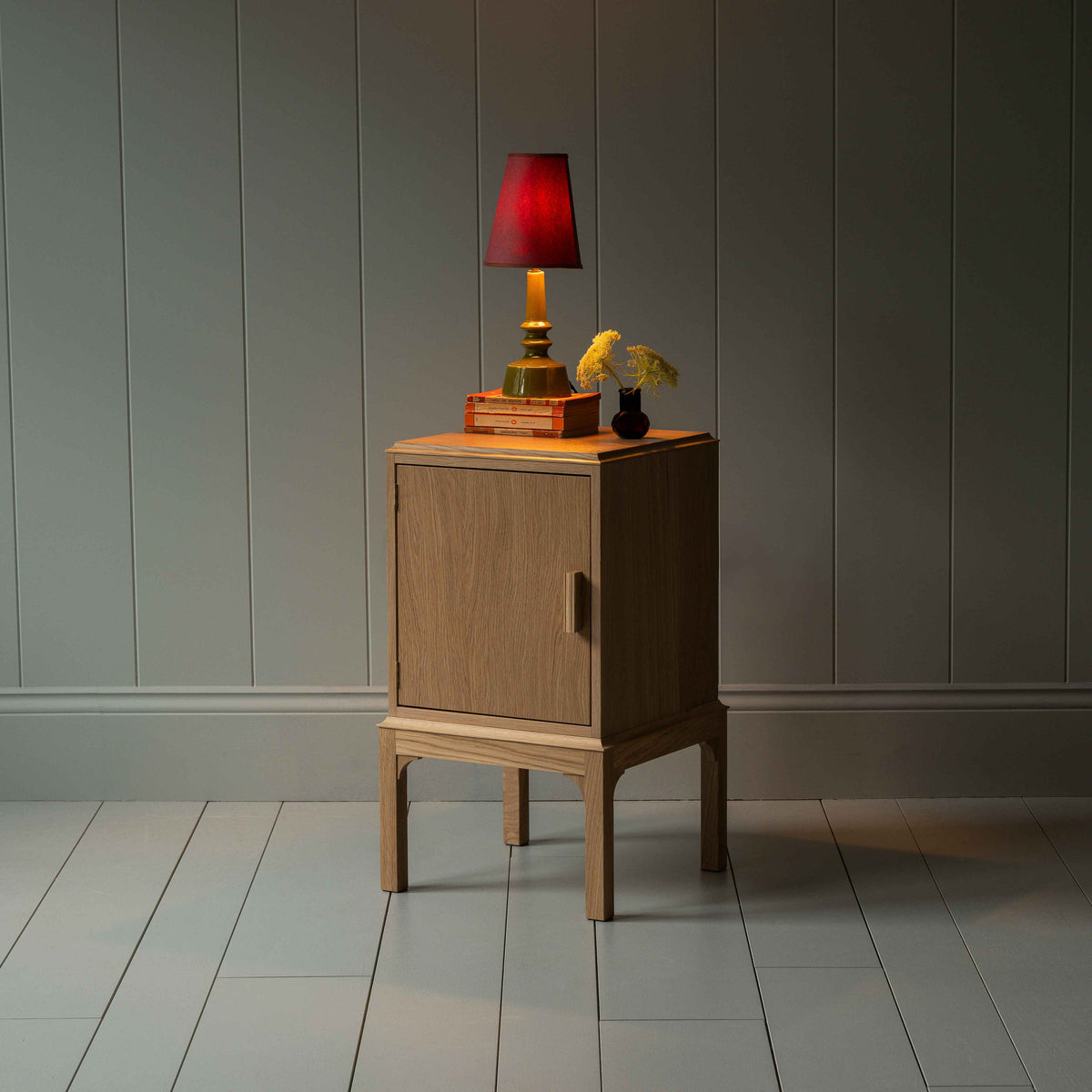 Bits and Bobs Bedside Table, Natural Oak, Left Hand Side Decorated With a table lamp and a small bottle of flowers - Nicola Harding