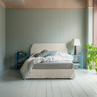 King-size upholstered bed by NiX in laidback linen dove with a kick pleat valance, featuring pillows and blankets, along with two side tables, one topped with a lamp