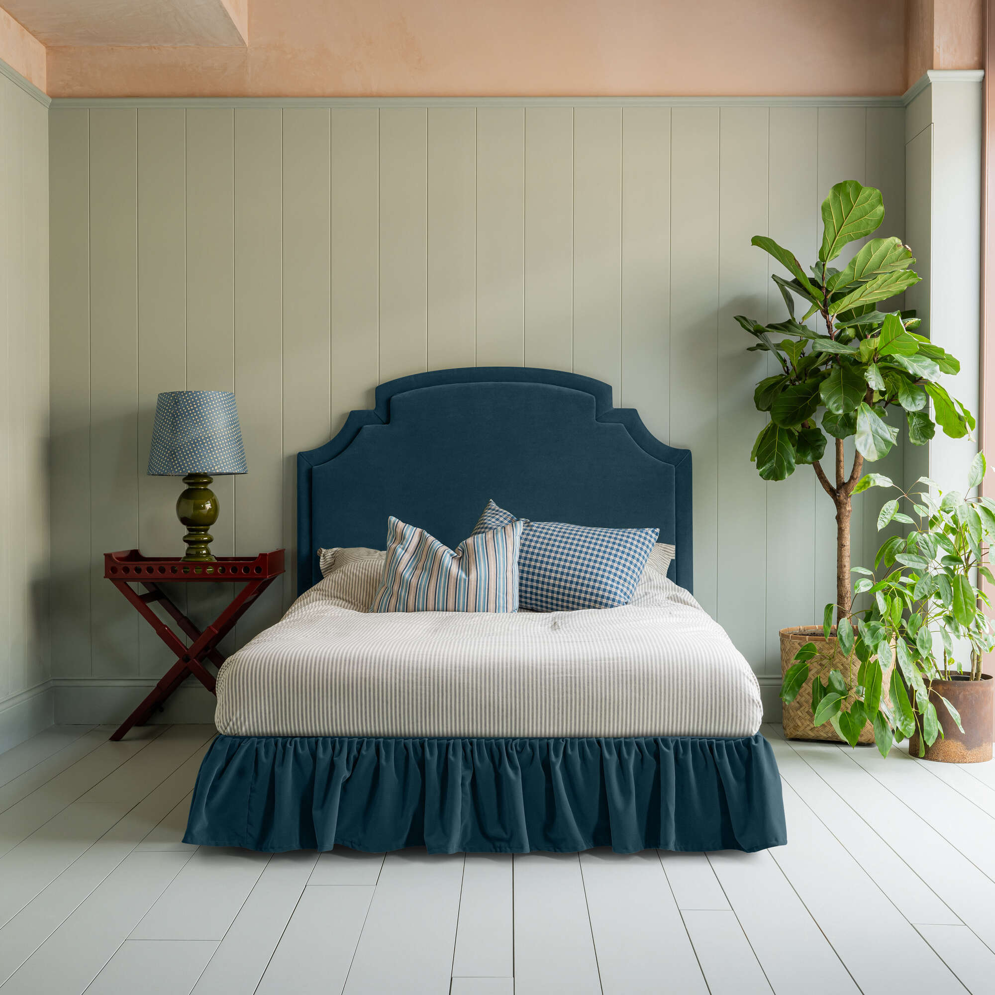  Seventh Heaven Upholstered Bed in Intelligent Velvet Aegean with Ruffled Valance, accompanied by a red side table with lamp, pillows, and a blanket - Designed by Nicola Harding 