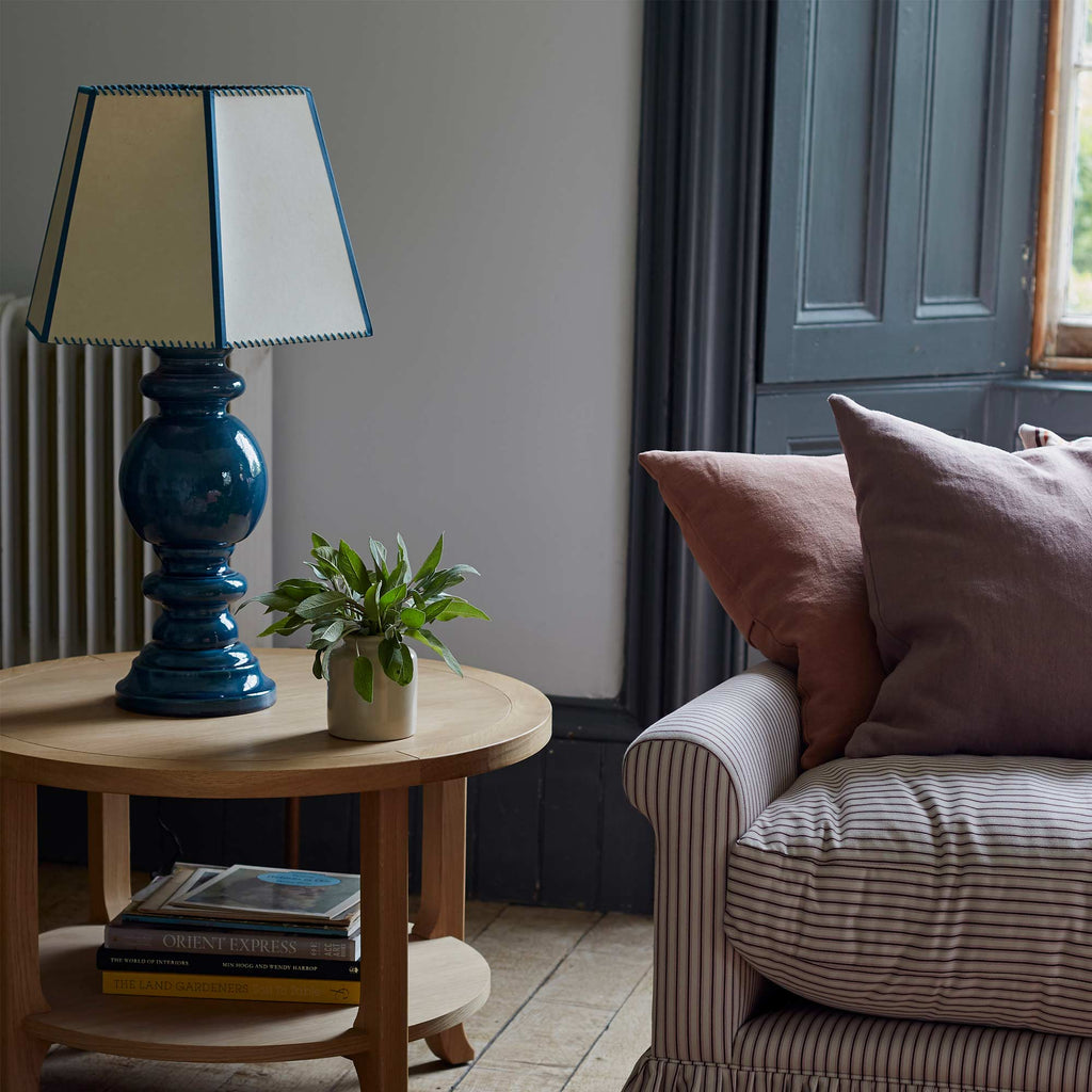  Hourglass Ceramic Table Lamp Base in Blue, With Townhouse Hexagonal Lamp Shade in Soft White with Peacock Blue Trim & Stitching On a Perk Coffee Table, Oiled Oak, With Couple of Books on the Bottom Shelf Right Next To NiX Sofa With Cushions - NiX By Nicola Harding & Co. 