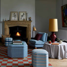 Orb Ceramic Table Lamp Base in Red, With Whimsical Tall Straight Empire Lamp Shade in Mustard with Antiqued Gold Trim, on a Side Table in the Living Room, With a Slipper Chair, 3 Ottomans Near a Fire Place - Nicola Harding