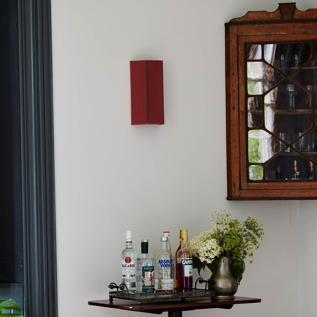  Stitch In Time Wall Light in Burgundy with Muted Pink Trim, Hanging Above a Bunch of Liquor Bottles and a Beautiful Plant – By NiX 