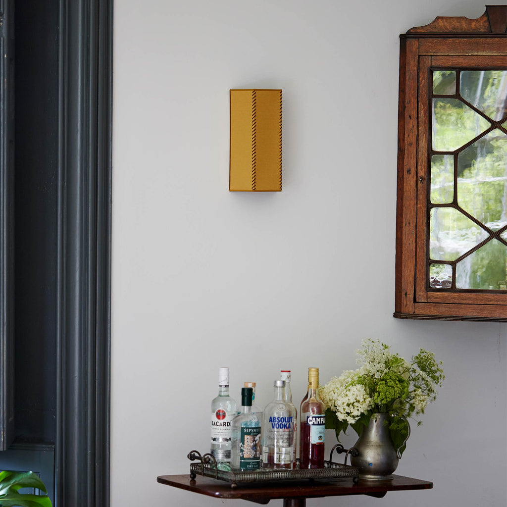  Stitch In Time Wall Light in Mustard with Antique Gold Trim, Hanging Above a Bunch of Liquor Bottles and a Beautiful Plant – By NiX 