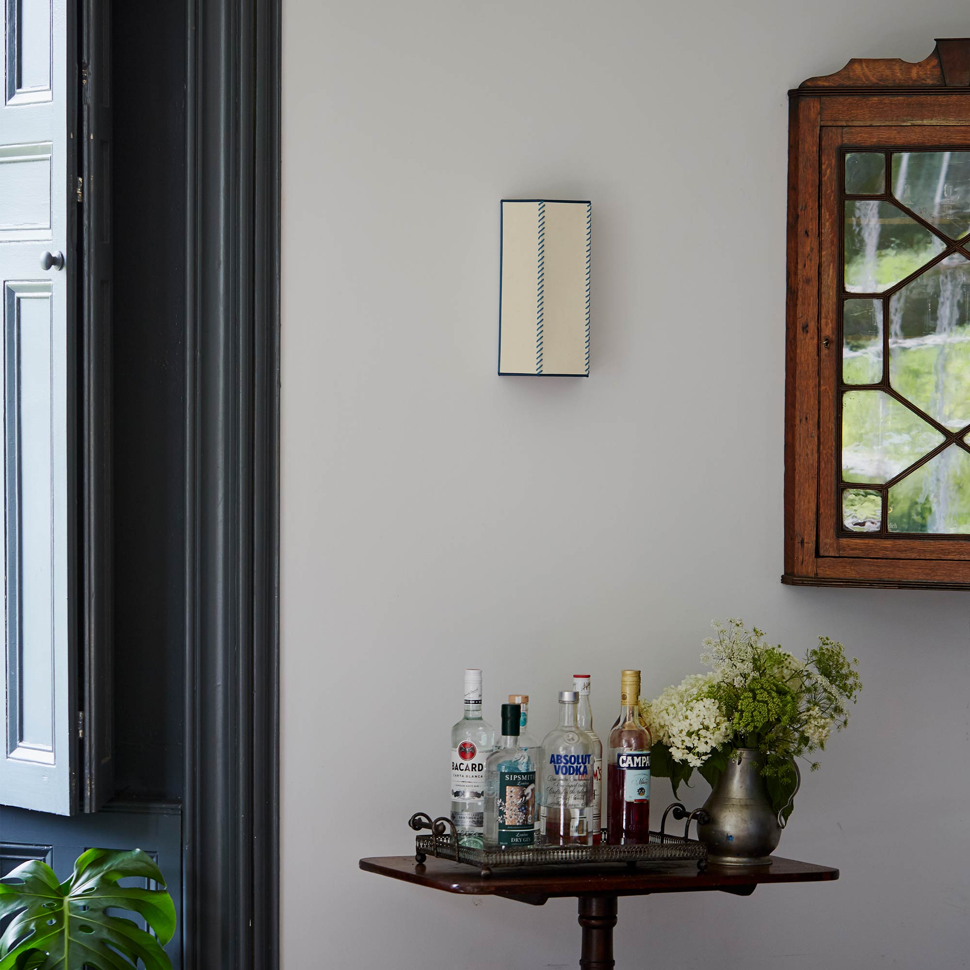  Stitch In Time Wall Light in Soft White with Peacock Blue Trim and Stitching, Hanging Above a Bunch of Liquor Bottles and a Beautiful Plant – By NiX 
